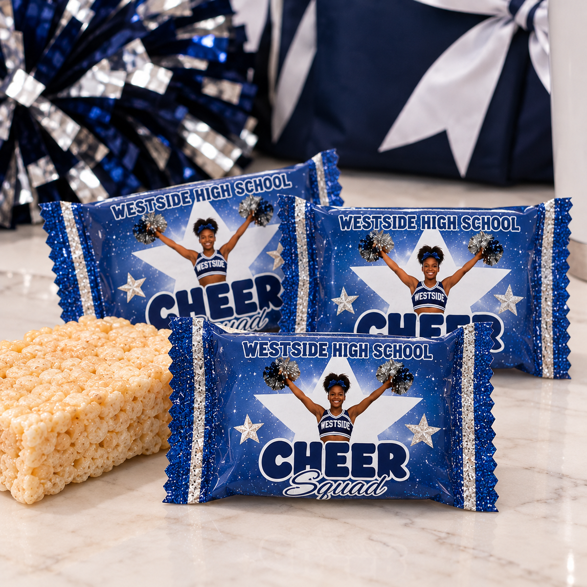 Three wrapped candy bars featuring a blue and silver glitter design with a cheerleader graphic and the text 'WESTSIDE HIGH SCHOOL CHEER Squads,' arranged on a marble surface next to a rice krisp treat. A blue and silver pom-pom head and a gift bag appear in the blurred background.